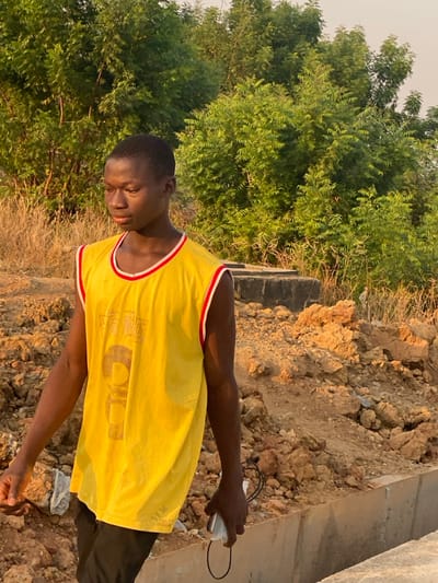A young man stands outside on uneven ground next to a concrete structure and leafy green trees. He wears a yellow and red sleeveless jersey, black pants, and holds a white object connected to a wire in his hand. He is looking down with his head tilted, possibly at something on the ground. The sunlight is bright, casting shadows across the scene. The surroundings appear to be a mix of natural and man-made elements.