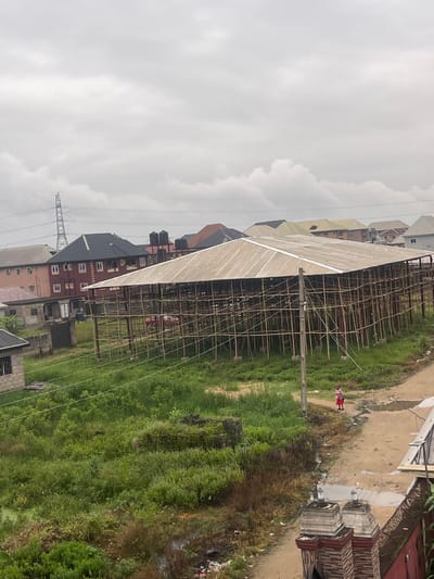 The image depicts an overcast day in Iyiowa Odekpa, Nigeria, with a large, open-sided structure covered by a corrugated metal roof supported by bamboo scaffolding occupying the center. In the background, various residential buildings with different roof colors and architectural styles are visible, along with a prominent electrical tower. To the left of the main structure, a multi-story brick building with a dark roof stands out. The ground surrounding the structure is covered in lush green vegetation, with some patches of muddy earth and puddles. A dirt path runs alongside the structure, on which a lone figure, a child in a pink shirt and red shorts, is walking away from the camera. In the foreground, part of a decorative gated entrance with stacked brick pillars and decorative tops is visible on the right.