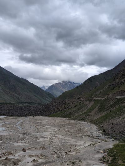 The media file depicts a dramatic mountain landscape in Darcha, India, under a cloudy sky. The primary subjects are the imposing, steep mountainsides and a dry riverbed or scree slope. The scene is characterized by rugged terrain, with the mountains covered in sparse vegetation and rocky outcrops.
The right side of the image features a wide, light-colored scree slope, heavily textured with rocks and pebbles of various sizes. Streaks of lighter and darker sediment suggest past water flow. The left side of the image is dominated by a dark, steep mountain face, also covered in rocks and patches of green vegetation. Further back, a glimpse of another mountain peak is visible, partially obscured by clouds, with snow-capped sections indicating high altitude.
The weather appears overcast, with thick, grey clouds filling the upper portion of the frame, creating a subdued and somewhat dramatic atmosphere. The lighting suggests it is daytime, but the clouds prevent direct sunlight. The overall mood is one of raw, natural beauty and the immense scale of the mountainous environment.
There are no visible people or specific objects that indicate a particular activity or event, other than the natural geological processes at play. The location cues, such as the mountain ranges and the general aridness of the scree slope, are consistent with a high-altitude, mountainous region like Darcha. No text is visible in the image.