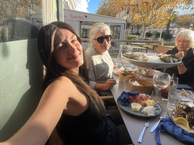 The image depicts an outdoor dining scene on a sunny day in La Grande-Motte, France, at a restaurant identifiable by a partial sign reading "es Caraïbes" (presumably Les Caraïbes). Three women are seated around a grey rectangular table. In the foreground, a young woman with long dark hair, wearing a dark top, is smiling directly at the camera, holding the device for the photograph. To her right, an older woman with short grey hair and sunglasses, wearing a light-colored top, is seated, looking towards the right side of the frame. Further to the right, another older woman with short light-colored hair, wearing a dark top, is engaging with a large, two-tiered silver platter laden with oysters on a bed of crushed ice.
The table holds various food items and beverages: the prominent oyster platter, an oval plate with peeled shrimp, lemon wedges, and a white sauce, and another plate with more oysters. Drinks include a glass of rosé wine, multiple clear tumblers, a clear bottle, and a carafe of red liquid. Woven baskets containing bread, silverware (forks and knives), blue napkins, and condiment dispensers are also present on the table. The background features trees with orange and yellow autumn foliage, a building structure, and a clear blue sky with some clouds, indicating strong natural light. The women appear to be engaged in a social meal.
