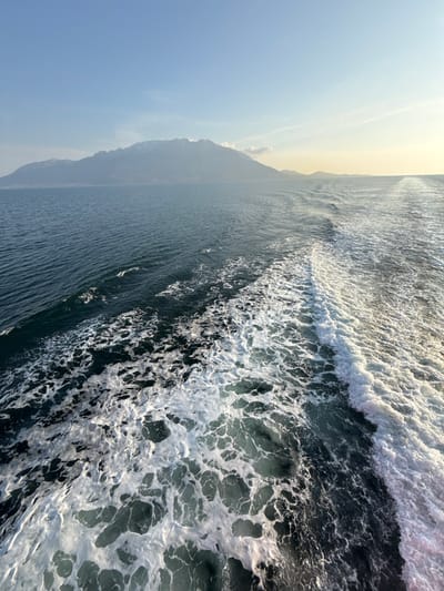 The image displays a wide marine landscape, depicting the frothy wake of a vessel against a backdrop of a mountainous landmass, situated in the vicinity of Palaiopoli, Greece. The foreground shows turbulent white foam and agitated dark blue-green water, extending diagonally from the lower left towards the upper right, indicating the movement of a boat. The water transitions to a deeper blue-green color further out, with visible ripples. A large, dark mountainous landmass dominates the background, stretching across the left and central frame. Its peaks exhibit faint patches of snow, and the entire landmass is softened by atmospheric haze. The sky above is clear and light blue, transitioning to a soft yellow glow on the right horizon, consistent with a low sun angle. No individuals, other vessels, or artificial structures are discernible within the frame, focusing solely on the natural seascape.