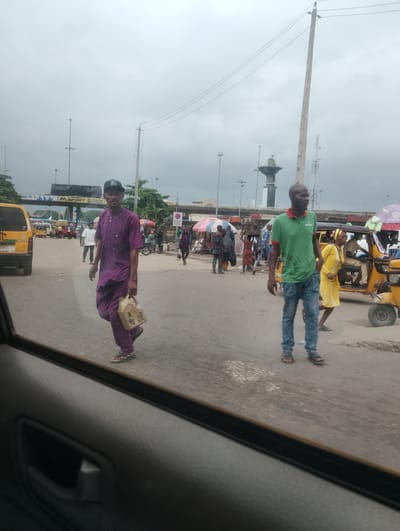 In this outdoor, medium-shot photograph, the scene depicts a street in Alagbede, Nigeria. The sky is overcast, suggesting a cloudy or potentially rainy day. The foreground is dominated by the interior of a vehicle, with a dark gray door panel and handle visible on the left. Reflected in the window, people are seen walking and gathered on a paved road.

Two men are prominently featured walking across the road. The man on the left is wearing a purple traditional outfit and a black baseball cap, carrying a light-colored plastic jerrycan. He is walking towards the viewer. The man on the right is wearing a green t-shirt with a red collar and blue jeans, with his hands in his pockets. He is walking away from the viewer.

In the background, there is a busy street scene with various vehicles, including a yellow van on the far left and a yellow auto-rickshaw to the right. Several other people are visible, some walking, others appearing to be vendors or passengers. Colorful umbrellas are set up, likely for market stalls. Infrastructure like utility poles with wires and what appears to be an overhead bridge or toll gate are also present. A tall, distinctive tower-like structure can be seen in the middle ground.

Text is visible on a sign in the background that reads "M.SPORT BET NOW ON". There are no clear indicators of the time of day, but the overcast conditions suggest it could be morning or afternoon. The overall impression is one of a typical day in a bustling Nigerian urban environment.