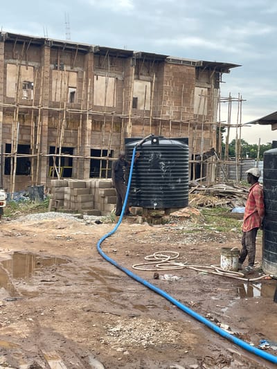 In Okinano, Nigeria, an unfinished multi-story building is under construction. The structure is made of concrete blocks with bamboo scaffolding supporting wooden planks on multiple levels, indicating ongoing work. In the foreground, two men are present. One man, wearing a dark shirt and pants, stands next to a large black water tank, holding a blue hose that extends into the tank. The second man, dressed in a red plaid shirt, dark pants, and a white cap, leans against another black water tank. The ground is muddy and uneven, with puddles reflecting the overcast sky, suggesting recent rain. Piles of concrete blocks and other construction debris are scattered around the site. White hoses are coiled on the ground near the second water tank.