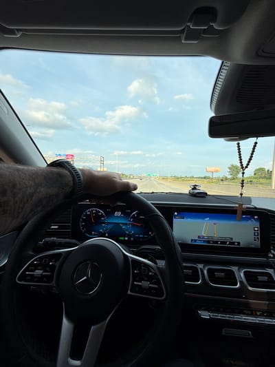 The image depicts the interior of a Mercedes-Benz vehicle, showing a driver's perspective on a multi-lane highway during daytime. The location is the Jane Addams Memorial Tollway (I-90) East, passing through or near the city of Des Plaines, United States of America, as indicated by the navigation screen and highway signs.
One person is present, visible by their right arm and hand resting on the steering wheel. The arm is hairy and bears a tattoo. A black smartwatch or fitness tracker is worn on the wrist.
Inside the vehicle, the digital dashboard displays a speed of 69 mph, an outside temperature of 81°F, and a time of 07:09. The fuel gauge indicates approximately half a tank, and the odometer/trip meter shows 437 miles. The transmission is in D9 (Drive, 9th gear), and a "READY" indicator is visible. The steering wheel, bearing the Mercedes-Benz logo, has various control buttons. To the right, a large infotainment screen displays a map showing "EAST Jane Addams Memorial Tollway" with live traffic information. A radar detector or similar device is mounted on the dashboard. Hanging from the rearview mirror is a rosary with wooden beads and a wooden cross. Below the infotainment screen are air vents and climate control buttons.
Outside the vehicle, a clear blue sky with scattered white clouds is visible. The multi-lane highway stretches into the distance, with other vehicles present. An overhead highway sign gantry with a blue "I-90" sign is visible. On the right side of the highway, a billboard partially displays "motel 6," and various trees and vegetation line the road.