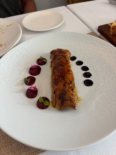 A close-up, high-angle shot shows a meticulously plated dish of food on a white, circular plate with a raised, embossed pattern. The main subject is a long, rectangular piece of golden-brown, glazed meat, possibly foie gras or a similar delicacy, positioned vertically in the center of the plate. To the left of the meat, five small, dark red spheres are arranged in a curve, each topped with a small green leaf and a delicate pink edible flower. To the right of the meat, five small, dark, glossy droplets, likely a sauce such as balsamic glaze, are arranged in a descending diagonal line. The plate rests on a white tablecloth. In the background, out of focus, are other elements of a dining setting, including a white side plate and a sliver of toast on a wooden board, suggesting a restaurant environment. The overall impression is one of fine dining and culinary artistry, likely in a restaurant in Carboneras, Spain.