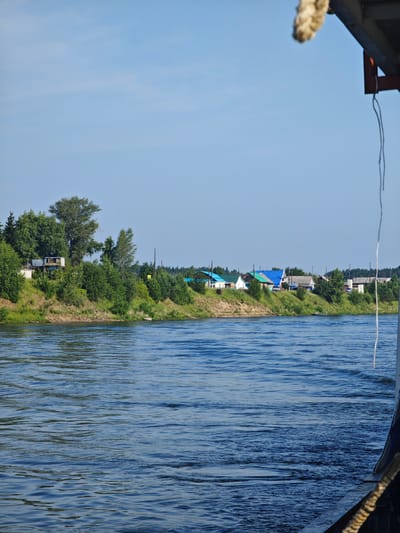 The image shows a view of the river in Galanino, Russia, from a boat. The water is blue and choppy, with ripples and waves. On the far bank, there is a line of trees and houses, with green roofs and white walls. The houses appear to be in a village, with power lines running between them. The sky is a clear blue, with a few wispy clouds. In the foreground, part of the boat is visible, with a thick rope tied to it. The overall impression is one of a peaceful, rural scene.