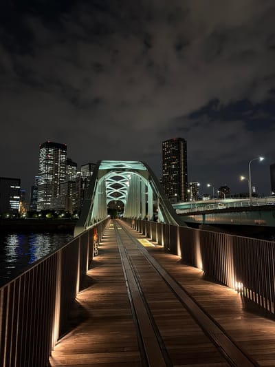 This media file captures a nighttime view of a modern pedestrian bridge in Koto, Japan. The bridge features a distinctive white, arched structure spanning across a body of water. The walkway of the bridge is made of wooden planks and is illuminated by strategically placed lights, casting long shadows and creating a warm, inviting ambiance.

On the left side of the image, several high-rise buildings are visible, their windows lit up, indicating a bustling urban environment. The sky is dark and cloudy, typical of a night scene. The reflection of city lights and the bridge's illumination can be seen on the water's surface, adding depth and visual interest to the scene.

There are no clear people visible, but there is an orange object that may be a bicycle or a cart parked on the walkway. The overall mood is serene and somewhat futuristic, highlighting the blend of modern architecture and nature at night. The scene suggests the bridge is a popular spot for evening strolls, offering a scenic perspective of the city. No discernible text is visible in the image.
