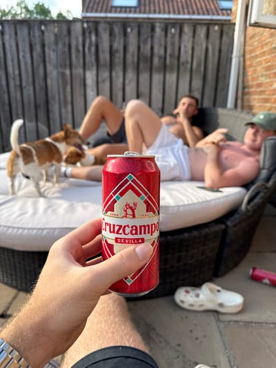 A man holds a red can of Cruzcampo Sevilla beer in the foreground. Two shirtless men recline on a white cushion in the background, with a small dog near their feet. A pair of white Crocs sits on the patio. The setting appears to be an outdoor patio area with a wooden fence and glimpse of a brick house.