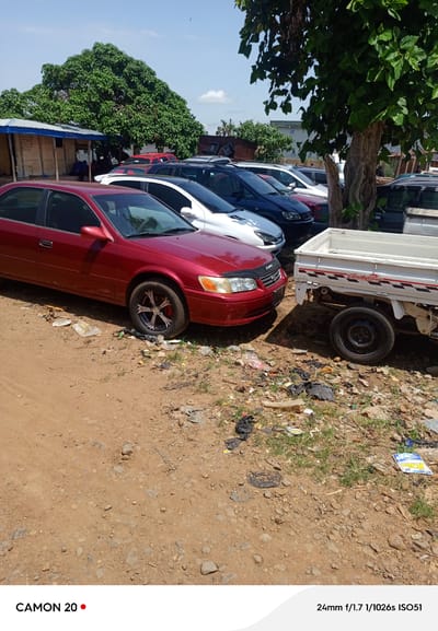 In Kafanchan, Nigeria, a red sedan is parked on a dirt road next to a white utility truck. The sedan is a Toyota Camry, and it has aftermarket rims and a black hood protector. In the background, there are other vehicles, a small building with a tin roof, and a large tree. The ground is covered in dirt, rocks, and litter. The sky is blue with a few clouds.