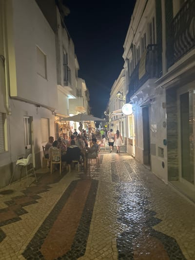 The image depicts a narrow cobblestone street in Lagos, Portugal, at night. The street is lined with buildings, some of which appear to be shops or restaurants with open-air seating. Several people are visible, mostly gathered around tables, dining and socializing. The cobblestones are wet, suggesting recent rain or cleaning, and reflect the ambient light. The overall atmosphere is lively and illuminated by artificial lighting from the establishments and street lamps.