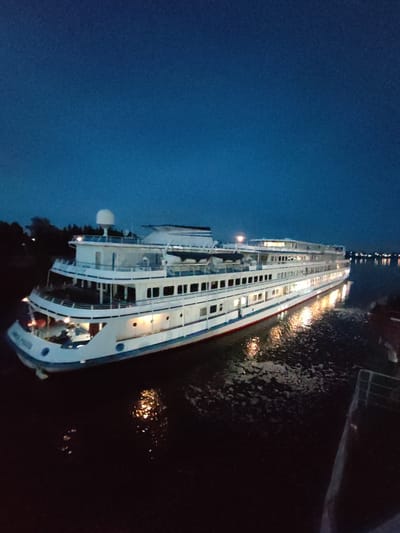 A large white passenger ship is docked on the dark, calm waters of the Sheksna River in Sheksna, Russia, during twilight. The ship, illuminated by interior lights and external spotlights, has multiple decks with rows of windows. The sky above is a deep, clear blue transitioning to darker hues. Reflections of the ship's lights shimmer on the water's surface. A dark landmass with trees is visible on the left bank of the river. The overall scene conveys a sense of peaceful repose as evening settles.