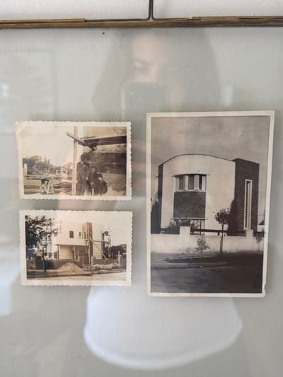 The image shows three black and white photographs, arranged in a glass frame with a metal border. The largest photograph is oriented horizontally and depicts a building with a curved facade and rectangular windows, likely a residence. Two smaller photographs, both with scalloped edges, are positioned below the larger one. The photograph on the left shows two adults and a child standing in what appears to be a garden or outdoor space, likely in a family portrait. The photograph on the right depicts the construction of the same building, with scaffolding visible. Vicente López, Argentina, is presumed as the location based on prior knowledge or implicit information.