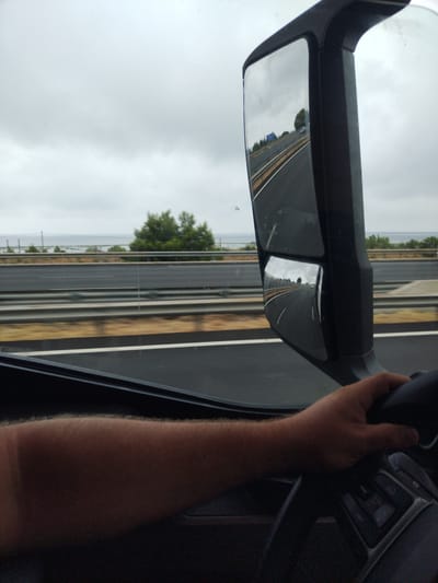 The image depicts a view from inside a vehicle, likely a truck, looking out a side window and into a side mirror. A person's arm and hand are visible, gripping a steering wheel. The scene outside shows a highway with multiple lanes, guardrails, and vegetation, including trees and bushes. In the distance, a body of water and a cloudy sky are visible. The side mirror reflects a portion of the highway, showing a vehicle and trees. The location is identified as Salses-le-Château, France, suggesting the highway is part of the French road network.