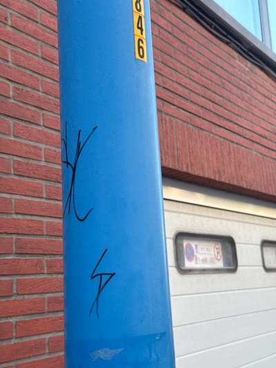 In Strombeek-Bever, Belgium, a bright blue cylindrical pole stands in front of a red brick building and a white garage door. A yellow sticker with the numbers "846" is affixed to the upper portion of the pole. Black graffiti marks are present on the pole, with a complex symbol resembling an abstract "X" or intertwining lines positioned in the middle, and a lightning bolt-like symbol further down. The garage door has two rectangular windows, one of which contains signage with a red circle and diagonal line crossed with a blue circle and red R, indicating parking restrictions. The environment suggests an outdoor urban or commercial setting.