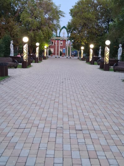 The image shows a paved pathway lined with benches and decorative lampposts in what appears to be a park or public square. In the background, there is a building with a red facade and a green arched structure above it. Several trees are visible, and the sky suggests it is either dawn or dusk. A few people are seated on one of the benches. The pathway is made of rectangular pavers in varying shades of beige and gray. The overall scene is one of a tranquil public space. Based on the provided information, this scene is located in Булаев қалалық әкімдігі, Kazakhstan.