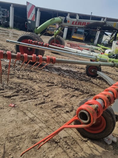 The image shows a hay rake implement on muddy ground in Ovcharovo, Bulgaria. The rake features multiple red tines attached to a metal frame with small wheels. A Claas tractor is partially visible in the background. The ground is covered in dirt and tire tracks.