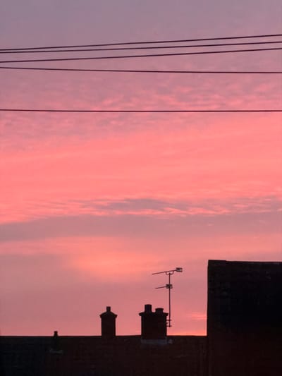 The image displays a silhouetted urban skyline in Bretton, United Kingdom, against a sky exhibiting a pink and light purple gradient with diffuse cloud formations, indicative of either sunrise or sunset. Four parallel overhead wires are positioned across the upper portion of the image. The lower part of the frame features the dark silhouettes of building rooftops. From left to right, a single chimney stack is visible, followed by a double-flue chimney structure. A television antenna, mounted on a pole, is situated on a rooftop to the right of the double-flue chimney. A substantial, dark, vertical building facade partially occupies the far right side of the frame, rising from the rooftop line. No human subjects, animals, or discernible activities are present.