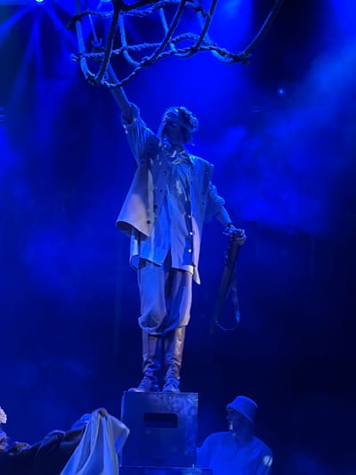This image depicts a theatrical performance or stage event under intense blue lighting in Novoshakhtinsk, Russia. A central figure, likely an actor, stands elevated on a dark, rectangular stage block. This individual is blindfolded, wearing a light-colored layered costume consisting of a shirt, an open vest with grommets, loose trousers, and dark tall boots. Their right arm is raised, grasping a dark, coarse rope net suspended above, while their left hand holds a dark, loop-ended strap. Partially visible in the lower frame are two other individuals: one on the left in dark clothing with a white cloth, and another on the right in a light-colored top and bucket hat, observing the central figure. The environment is characterized by strong blue stage lighting and visible atmospheric haze.