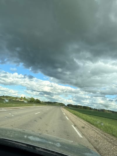The image depicts a road in Beržiniai, Lithuania, as seen from inside a vehicle. The road is paved, straight, and marked with white lines. To the right of the road is a grassy verge, then a gravel area, and finally a dense line of trees. The sky occupies a large portion of the frame, featuring a dramatic cloudscape with both dark, ominous storm clouds and patches of blue sky. The foreground includes part of the vehicle's interior, specifically the side window frame with some raindrops on it. The focus is sharpest on the road and its immediate surroundings.