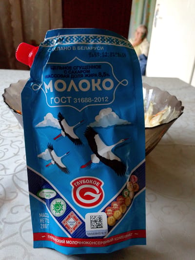 The image shows a close-up of a blue pouch of condensed milk with the brand name "МОЛОКО" (Milk) prominently displayed. The pouch features illustrations of storks in flight against a blue sky with white clouds. Text on the pouch indicates it is made in Belarus, contains sugar, has a fat content of 8.5%, and complies with GOST 31688-2012. There are also various stamps and seals on the pouch, including one that says "ЛИДЕР 2023" (Leader 2023) and a "HALAL" certification. A red cap is visible on the top left of the pouch.

In the background, a blurred image of an elderly woman is visible, possibly in a home setting. She is wearing a light-colored top and has her hand near her face. The background appears to be indoors, with a door and window visible. The overall scene suggests a casual, domestic setting, with the pouch of condensed milk being the primary focus. The lighting indicates it might be daytime.