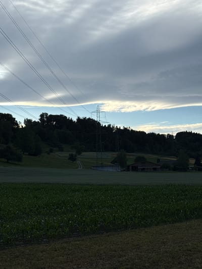 The image depicts a rural landscape in Lufingen, Switzerland. In the foreground, a field of young corn plants stretches across the frame, with dry grass visible at the very bottom. Behind the cornfield, a larger, darker green field, possibly a different crop or pasture, is visible. In the mid-ground, a gently sloping hill is covered in grass, with a dirt path winding through it. Scattered trees and bushes dot the hillside, and a small cluster of buildings, including a barn-like structure, is situated on the right side of the hill. A prominent set of power lines, supported by a large transmission tower on the right, traverses the scene diagonally from the top left towards the mid-right. The background is dominated by a dense line of dark green trees forming a forest that crowns the hill. The sky above is dramatic, with thick, layered clouds ranging from dark gray to lighter shades, interspersed with streaks of bright blue and hints of white light, suggesting either early morning or late evening. The overall impression is a serene, agricultural setting under a dynamic sky.