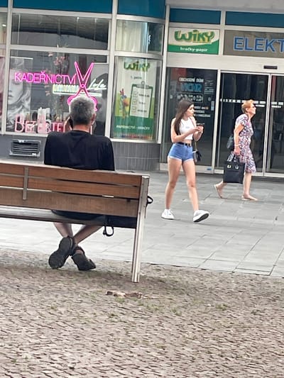 In Brno, Czechia, on a paved sidewalk outside commercial establishments, a man with gray hair sits on a wooden park bench facing away from the camera, talking on a mobile phone. He is wearing a black t-shirt and shorts, with his legs crossed and feet wearing dark-colored shoes. To his right, a young woman in a white crop top and denim shorts walks by, looking at her phone and carrying a black purse. Further to the right, an older woman with short blonde hair walks away from the camera, wearing a floral print dress and carrying a black handbag. Behind them, storefronts display signage, including "KADERNÍCTVÍ LALA" with neon pink scissors and "WIKY" with text below indicating toys and paper. The ground is a mix of cobblestones and lighter paved tiles.