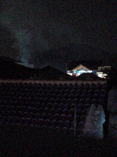 This image depicts a nighttime scene in Bojongherang, Indonesia, characterized by low light and significant digital noise. The foreground is dominated by a dark, textured surface consistent with a tiled roof, featuring multiple rows of overlapping, convex tiles extending from left to right across the lower half of the frame. On the far right edge of the foreground, a faint, vertical, lighter-colored object, possibly a structural element or wall, is partially visible.
In the mid-ground, beyond the tiled roof, several structures are present. A prominent building in the center features a light-colored, possibly gabled, roof and is illuminated by bright artificial lights, casting a strong bluish-white glow. These lights appear to originate from within or directly on the building, illuminating its facade and creating a stark contrast against the surrounding darkness. Additional, less distinct structures are visible around this illuminated building, appearing darker and less defined.
The background consists of dark, indistinct shapes, possibly hills or distant buildings, further obscured by the extreme low light and image noise. No discernible individuals, specific actions, or interactions are visible within the frame. The overall environment suggests an urban or semi-urban setting at night, with artificial illumination concentrated on one or more buildings.