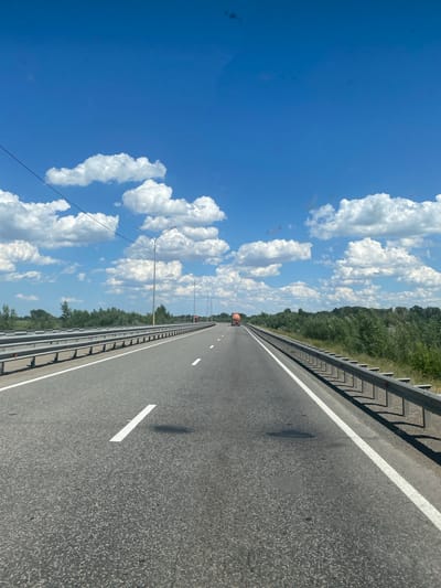 The image shows a road with a blue sky and fluffy white clouds. The road is asphalt and has white lane markings. There is a metal guardrail on the right side of the road, and a line of trees and vegetation on the left. In the distance, there is a red truck. The image appears to be taken on a sunny day, likely in the afternoon, given the bright light and clear sky. The overall scene suggests a highway or a large road in a rural or semi-rural area. There are no people or text visible in the image.