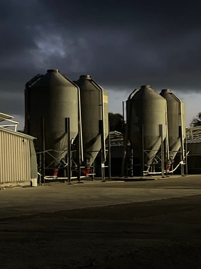 Four cylindrical tanks are stacked vertically against a cloudy night sky. Each tank has a conical bottom and a corrugated surface. Metal scaffolding supports the tanks, with glimpses of red machinery or fixtures visible. A concrete or paved surface stretches to the right, while a staircase leading up to the tanks is seen in the foreground. The image is dimly lit, casting long shadows.