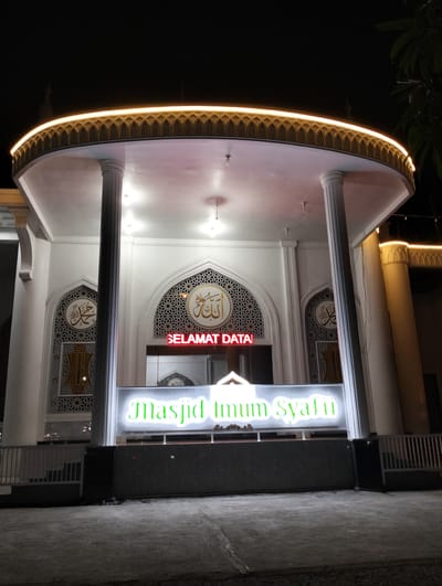 This is a nighttime, eye-level, outdoor, medium shot of the entrance to a mosque in Blangmetimu, Indonesia. The mosque is illuminated by a warm yellow light that outlines the curved roofline and runs along the top of the building. The facade is white, with decorative arches above the entrance. Above the main entrance, red neon lights spell out "SELAMAT DATA," which translates to "Welcome." Below this, a large, illuminated sign in green cursive reads "Masjid Imum Syafi'i." Two large white columns flank the entrance. Arabic calligraphy in gold is visible within decorative archways on either side of the main entrance. A white metal fence runs along the front of the property, and the ground in front is paved with concrete. The overall impression is one of a welcoming and well-lit place of worship.