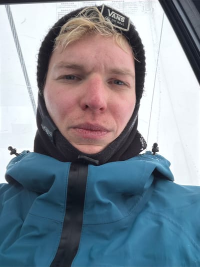 A selfie shows a fair-skinned person wearing a black Vans beanie and a blue winter jacket with a black stripe across the chest. They are also wearing a black neck warmer. The person has light blonde hair and blue eyes. The background appears to be a window with a metal cable running across it, possibly in a gondola or ski lift. The lighting is soft and diffused, suggesting an overcast day.