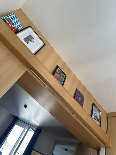 A low-angle interior shot shows a section of built-in wooden shelving against a white wall. The shelves are adorned with various framed pictures, a book, and an artwork. The lighting suggests daylight entering from a window partially visible in the lower right corner. The shelves are constructed of a light-colored wood. The perspective is from below, looking up at the shelves which are mounted along the wall and angling toward an opening to another room, where a window with blue curtains is visible. Saint-Ouen-sur-Seine, France is not directly discernible from the image content.