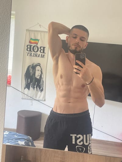 The image, taken in Tihomir, Bulgaria, shows a man with short brown hair and a beard posing in front of a mirror. He is shirtless and wearing black shorts with white lettering. His left arm is raised behind his head, and he holds a smartphone in his right hand, taking a selfie. In the background, a banner featuring Bob Marley hangs on the wall, alongside a large dark-colored ottoman and a portion of a bed with blue and white star-patterned bedding. The scene appears to be an indoor setting, likely a bedroom.