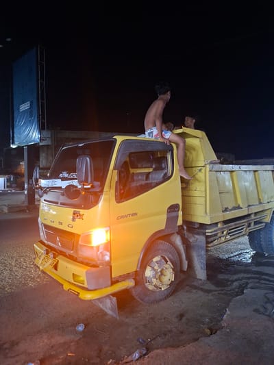 An outdoor, nighttime photograph shows a yellow Mitsubishi Canter Fuso dump truck overturned on its left side on a muddy or unpaved road. The truck's headlights are illuminated, casting a warm orange glow on the immediate surroundings.
Two shirtless individuals, likely young males, are visible interacting with the overturned vehicle. One person, wearing patterned shorts, is positioned with their legs extending upwards from the truck's bed, suggesting they are either inside the cargo area or climbing on it. A second person is partially visible behind them, peering over the edge of the truck bed.
The ground is wet and uneven, covered in mud and dirt, with scattered debris such as plastic bottles and wrappers near the truck's front bumper. In the background, to the left, a dark blue tarp or billboard structure with metal railings is partially visible against the dark night sky.
Visible text on the truck includes "CANTER," "FUSO," and "SUPER HDX 6.6" on the cab. A sticker on the side window reads "PUTRI ARDHANA." The license plate "RL 8750 DB" is clearly seen, along with "06.28," likely an expiry date. The blue structure in the background displays two phone numbers: "0813 7233 4001" and "0813 7120 7752." The scene depicts the aftermath of an incident involving the truck in Keudegeudong, Indonesia.