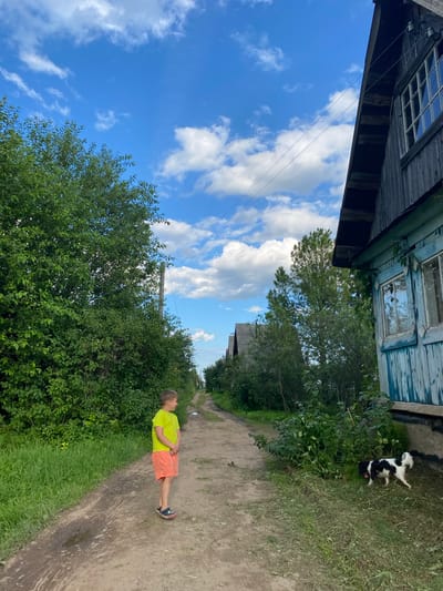 A young boy in a yellow t-shirt and orange shorts lies on a dirt road, his eyes closed and hands clasped over his stomach. He appears to be sleeping or resting in the shade of lush green bushes. The road is flanked by dense vegetation and a weathered, wooden building with peeling blue paint. In the background, a small black and white dog runs across a grassy area next to the building. The sky is a vibrant blue with scattered white clouds, suggesting it is daytime and the weather is pleasant. The scene has a rustic, rural feel, consistent with the location context of Novyi Ural, Russia. There is no visible text in the image.