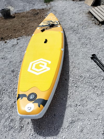 A bright yellow inflatable stand-up paddleboard lies on a gravel surface. The board is oriented lengthwise from the bottom right to the top left of the frame. It has a textured yellow deck with a white geometric logo in the center and images of black dolphins near the front. A black coiled leash is attached to the board. Two black paddle shafts with handles are visible to the right of the board, lying on the gravel. To the left of the board, in the background, is a dark gray bucket and a patch of dirt with some sparse vegetation. In the upper right corner, a wooden pallet is partially visible. The scene appears to be outdoors on a sunny day, indicated by the strong shadow cast by the paddleboard. The overall impression is one of preparation for or a break from water activities. The visible text on the board is a logo and "Inflatable Stand Up Paddle Board". The location is Kudashivka, Ukraine.
