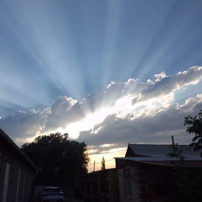 This is a landscape photograph taken in Almaluu, Kyrgyzstan, likely during the late afternoon or early evening. The primary subject is the sky, which is dominated by dramatic sunbeams (crepuscular rays) radiating from behind a large band of clouds. The sun itself is obscured, but its bright light pierces through gaps in the clouds, creating a striking visual effect. The sky is a gradient of blues, with the sunlit clouds appearing white and bright, while the clouds lower down and on the edges are darker, suggesting their thickness and the approaching end of the day.

In the foreground and midground, there are elements of a rural or suburban setting. To the left, a dark silhouette of a building with a grey, textured exterior and white doors or windows is visible. Further to the right, the dark silhouettes of other structures, including a building with a corrugated metal roof and brickwork, are present. A tall, slender tree stands out in the midground, its dark foliage contrasting with the bright sky. A white car is parked near the building on the left.

The overall mood of the image is serene and awe-inspiring, due to the dramatic interplay of light and shadow in the sky. The crepuscular rays create a sense of wonder and grandeur, while the darkened silhouettes of the houses and trees anchor the scene in a terrestrial reality. The weather appears to be partly cloudy, with the sun breaking through. No visible text is present in the image.