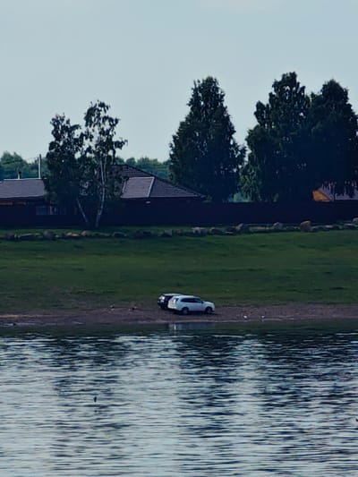 The image captures a serene waterscape with a shoreline dotted with two parked cars. The larger, white car, possibly a SUV, is positioned on the right side of the frame, while a dark-colored car is situated to its left. The cars are parked on a strip of dry land adjacent to a grassy embankment. This embankment leads up to a residential area, characterized by dark-paneled houses with prominent brown roofs. A line of large, rough-hewn stones marks the transition between the embankment and the residential area. The background is filled with dense foliage, dominated by tall, dark green trees, suggesting a rural or suburban setting. The sky above is a pale, cloudless blue. The foreground of the image is the surface of a body of water, showing subtle ripples and reflections, giving the impression of a calm day. While the specific location is identified as Erkalovo, Russia, the image itself provides no further context about human activity beyond the parked vehicles.