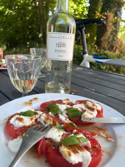 In Bièvres, France, a close-up shot captures a table setting outdoors, likely on a patio or in a garden, with lush green trees and foliage in the background. A white wine bottle, labeled "Domaine de la Tour du Bon, BANDOL," stands upright. Next to it are two clear wine glasses, one partially filled with white wine. In the foreground, a white plate displays a Caprese salad composed of sliced tomatoes, fresh mozzarella, and basil leaves, drizzled with a dressing that appears to include mustard seeds. A fork rests on the left side of the plate, and a knife is positioned on the right, suggesting an ongoing meal. In the blurred background, a bicycle is visible, hinting at a recreational setting. The lighting indicates it is daytime, with the sun filtering through the trees.