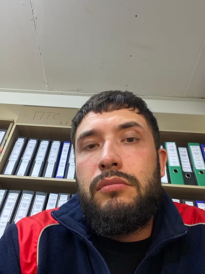 The image is a selfie taken by a man with dark hair and a beard, wearing a dark blue jacket with red accents. He is looking directly at the camera with a neutral expression. Behind him, a shelf filled with binders is visible. The shelf has some writing in red marker that appears to read "ПТС 4.1". The environment appears to be an office or storage area. The ceiling above is white and shows some signs of wear. The man is positioned in the foreground, with the shelves and binders slightly out of focus in the background. The lighting is bright, suggesting indoor illumination. The image's context places it in Luzhitsy, Russia.