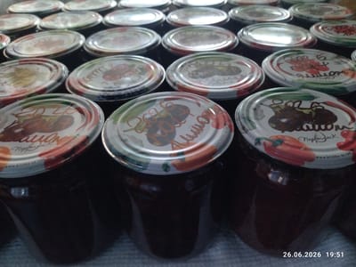 The image displays a close-up, overhead view of numerous jars of preserves, likely homemade, arranged in a grid. The jars have metal lids adorned with colorful illustrations of fruits and vegetables, and some also feature text. The preserves themselves are dark in color, appearing to be jam or jelly. The lids have a metallic sheen, reflecting the light source.
Several jars prominently feature the year "2026" written in an orange script, suggesting they were made in that year. One lid has the Cyrillic text "Нервбаст" and "www.krishka-ua.com", indicating a brand or origin. Another lid shows a drawing of peppers. The overall scene suggests a food preservation activity, likely taking place in a home or a small production setting.
The lighting is somewhat dim, with reflections on the jar lids indicating an indoor setting. The focus is sharpest on the jars in the foreground and middle ground, with those in the background becoming progressively blurred. The lower right corner of the image displays a timestamp: "26.06.2026 19:51," suggesting the photograph was taken on June 26, 2026, at 7:51 PM. The mention of Zavodske, Ukraine, provides a geographical context for this scene. There are no people visible in the image.
