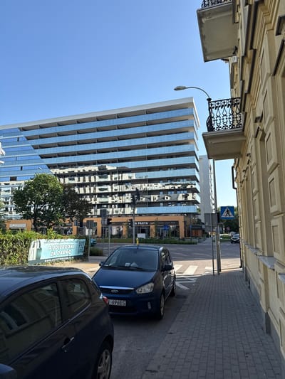 This image shows a street scene in Białystok, Poland. The perspective is from a low angle, looking up and towards a modern glass-fronted building. The building's facade reflects the blue sky and other surrounding structures, including an older, ornate building on the left and a street with cars and a pedestrian crossing in the distance.
In the foreground, two cars are parked along the street: a dark-colored car on the right and a blue car behind it. A street lamp arcs over the sidewalk. A large, leafy tree stands near the base of the modern building, partially obscuring some signage.
Visible text on a sign reads "AUDIOBOOKI ZA DARMO" which translates to "Audiobooks for free," and a website address "WWW.POZYTYWNEAUDIO10.PL". Other visible text includes "PLAZA", "PASTA", "Centro", "pizza wino Steki", and a license plate "696ES".
The scene appears to be a bright, sunny day, suggested by the clear blue sky and the sharp shadows cast by the buildings and cars. The overall impression is a blend of old and new architecture in an urban environment. The focus is on the buildings, cars, and street infrastructure, with no people clearly visible.