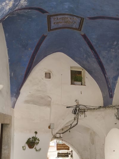 The image captures an arched passageway in Sperlonga, Italy, with a fresco on the ceiling depicting a star-filled blue sky. A sign within a rectangular frame on the ceiling reads "PIAZZETTA DELLA CONCORDIA" and a date "2 MARZO 1915". White, curved walls form the structure of the passage. To the left, a hanging potted plant with green foliage is affixed to the wall. A small square opening is visible on the upper left wall. Further down the passageway, a window with a potted plant in a basket can be seen. Electrical wires and conduits are strung across the ceiling of the passageway. Two wooden signs are visible hanging from the ceiling, one reading "MICHELANGELO IMMOBILIARE" with an arrow pointing right, and the other "holidaycasa GESTIONE CASE VACANZA". The lighting suggests it is daytime, with natural light filtering into the passage.