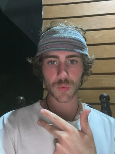 A male subject is depicted in a medium-shot portrait, located in Baan Nai Wok, Thailand. He is a young adult with light brown, wavy hair, partially contained by a horizontally striped knitted headband in shades of light blue, purple, pink, and grey. His blue eyes are directed towards the camera. The subject has a prominent mustache and his cheeks and nose exhibit a flushed appearance. He is wearing a white crew-neck t-shirt and a thin, dark cord necklace. His right hand is raised to chest level, displaying a "shaka" gesture with the thumb and pinky finger extended, and the middle three fingers curled. The skin on his hand exhibits a sheen, consistent with moisture or reflective residue. The background features a light brown wooden slatted wall on the right side of the frame, running horizontally. The left side of the background is dark and indistinct, with a partial view of a dark, rounded object featuring a reflective top surface in the lower left quadrant.