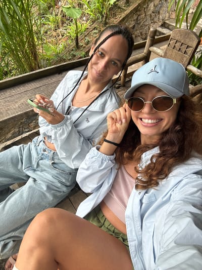 Two women are posing for a picture outside in Yên Minh Commune, Vietnam. The woman on the left is wearing a light blue jacket, light blue jeans, and her hair is in braids. She is holding a green phone. The woman on the right is wearing a light blue cap, sunglasses, and a light blue jacket. They are sitting on a wooden bench. Green plants are visible in the background.