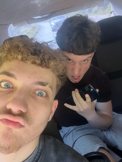 The image shows two young men inside a vehicle, possibly a car. The man in the foreground, on the left, has curly blond hair and light-colored eyes. He is looking directly at the camera with his mouth slightly open. The man behind him, on the right, has short, dark hair and is making an angry or surprised facial expression, with his mouth open and teeth visible. He is holding a dark-colored smartphone in his right hand. Both individuals are wearing casual clothing. The interior of the vehicle is visible, with a headliner in the background that has small reflective elements. The rear window of the vehicle is also visible, showing a blurred outdoor scene with trees and what appears to be a building or structure. The image is taken from a low angle. The specific location in Vlahovo, Bulgaria, cannot be determined from the visual information provided in the image.