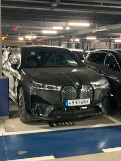 The image shows a black BMW iX parked in an indoor parking garage. The car is facing forward, and its license plate, "4898 MHK," is visible. The parking garage appears to be well-lit with fluorescent lights on the ceiling. There are other cars parked nearby, including a dark-colored SUV to the right of the BMW. The floor of the parking garage is concrete with blue and white painted markings. The overall scene is a typical indoor parking facility. The image was taken in Manises, Spain.