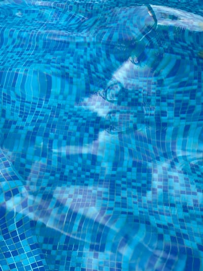 The image shows the bottom of a swimming pool covered in blue tiles. The water's surface creates rippling distortions and reflections that obscure the tile pattern. Reflected in the water is a face. The location is unknown, and there are no details indicating it is San Francisco Lachigoló, Mexico.