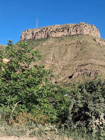 This is a vertically oriented, eye-level medium shot of a rugged mountain landscape in Shatin, Armenia. The scene is dominated by a massive, striated rock face on the left, which appears to be a cliff or a steep hillside. The rock face is a light brown and tan color, with distinct horizontal layers and some vegetation clinging to its surface. To the right of the rock face, the terrain transitions to a grassy, undulating slope.
In the foreground, lush green foliage from trees and bushes obscures part of the lower section of the image. Some of these plants appear to have small, round fruits or berries. Further into the foliage, there are hints of man-made structures, including what looks like a small, dark-colored building or shed with a metal roof, and possibly some other debris. There is also a tall, thin metal structure with crossbars visible on the rock face, likely a communication tower or part of a power line.
The sky is a clear, bright blue, suggesting a sunny day. The lighting indicates it is likely daytime, possibly late morning or early afternoon, given the shadows cast. The overall impression is one of a wild, somewhat remote natural environment with subtle signs of human presence. There is no discernible text in the image.