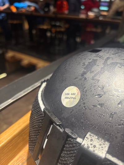 The image displays a wet, dark gray or black ski helmet positioned in the foreground, resting on a light brown wooden surface. The helmet's exterior is uniformly covered with numerous small water droplets, suggesting recent exposure to moisture. A circular, faded yellow-white sticker, approximately 4 cm in diameter, is affixed to the helmet's side, featuring the black capital text "YOU ARE AMAZING". Visible below the helmet's rim are elements of a black fabric strap with plastic buckles and a narrow edge of white interior padding. The background is significantly out of focus, revealing an indoor commercial setting consistent with a restaurant, bar, or ski lodge in Sankt Anton am Arlberg, Austria. A long, dark wooden counter or table extends horizontally across the mid-ground. Behind this counter, several blurred figures of individuals are discernible, some wearing outerwear including blue and red jackets. The lighting is ambient and somewhat dim. No specific actions or interactions are clearly visible among the background figures.